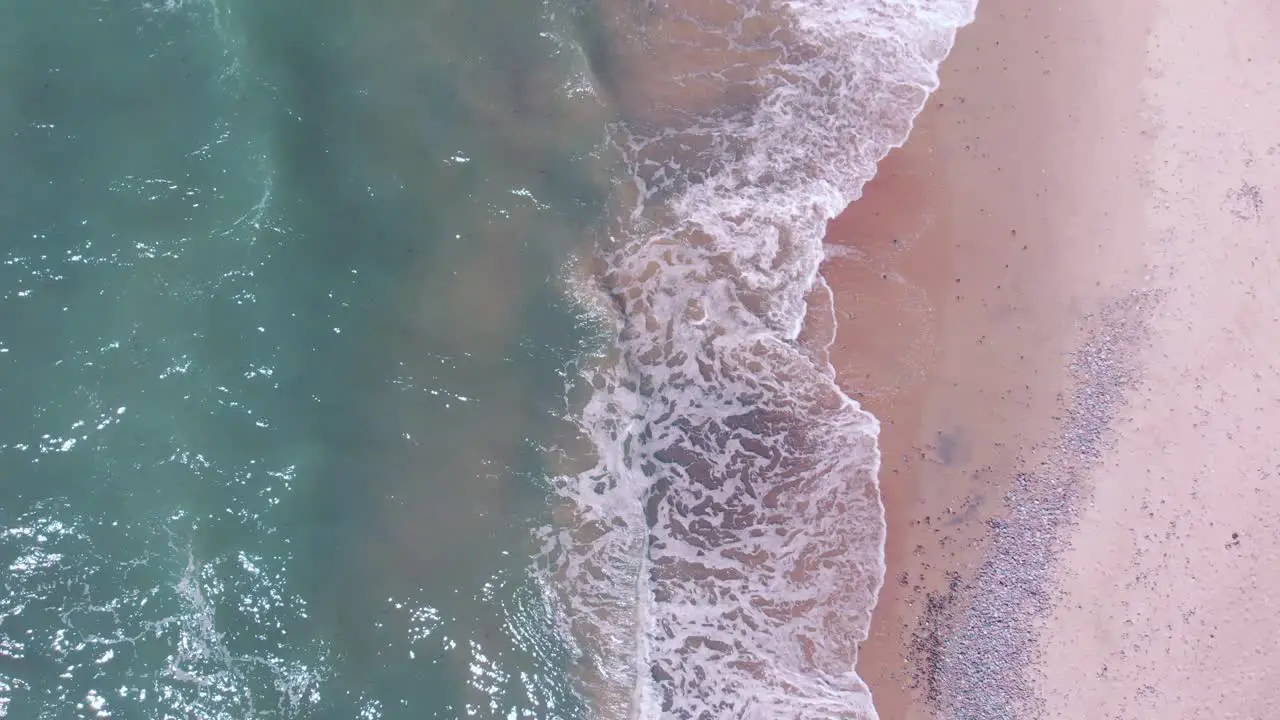 Top aerial view of ocean waves splash against pink tropical beach on summer day zooming out