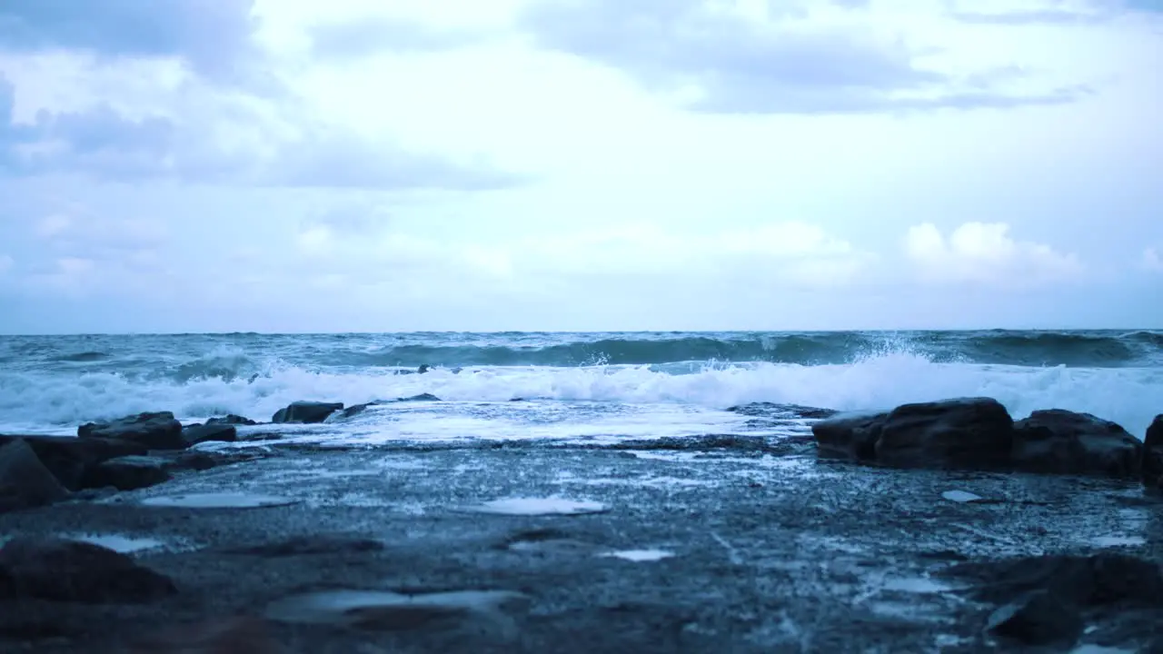 Crabs eye view of small cold waves crashing against a rocky jetti that stretches into the sea as the setting sun leaves a blue tint in the sky water and clouds