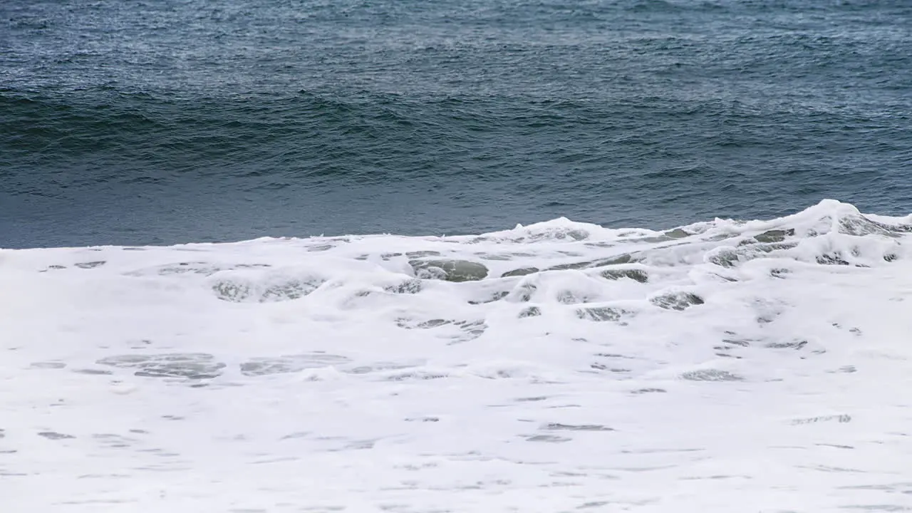 Small wave forming and crashing in slow motion in the shore with a deep dark blue ocean behind it and white water spilling in high speed camera 120fps 4K footage