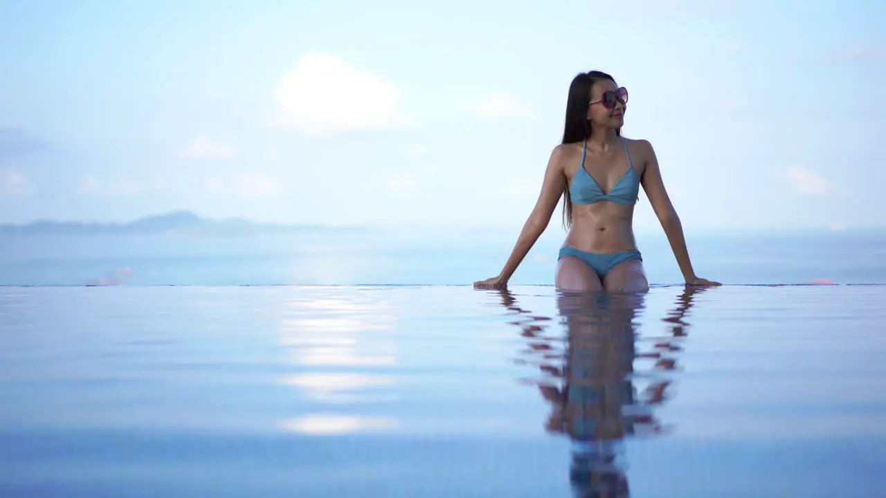 Pretty fit young Asian woman in a blue bikini sits on the edge of a resort infinity pool enjoying the warm tropical sun