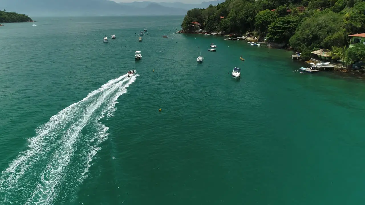 Aerial drone shot of lots of speedboat passing through moored boats near the coastline of a tropical forest with dense vegetation and few constructions