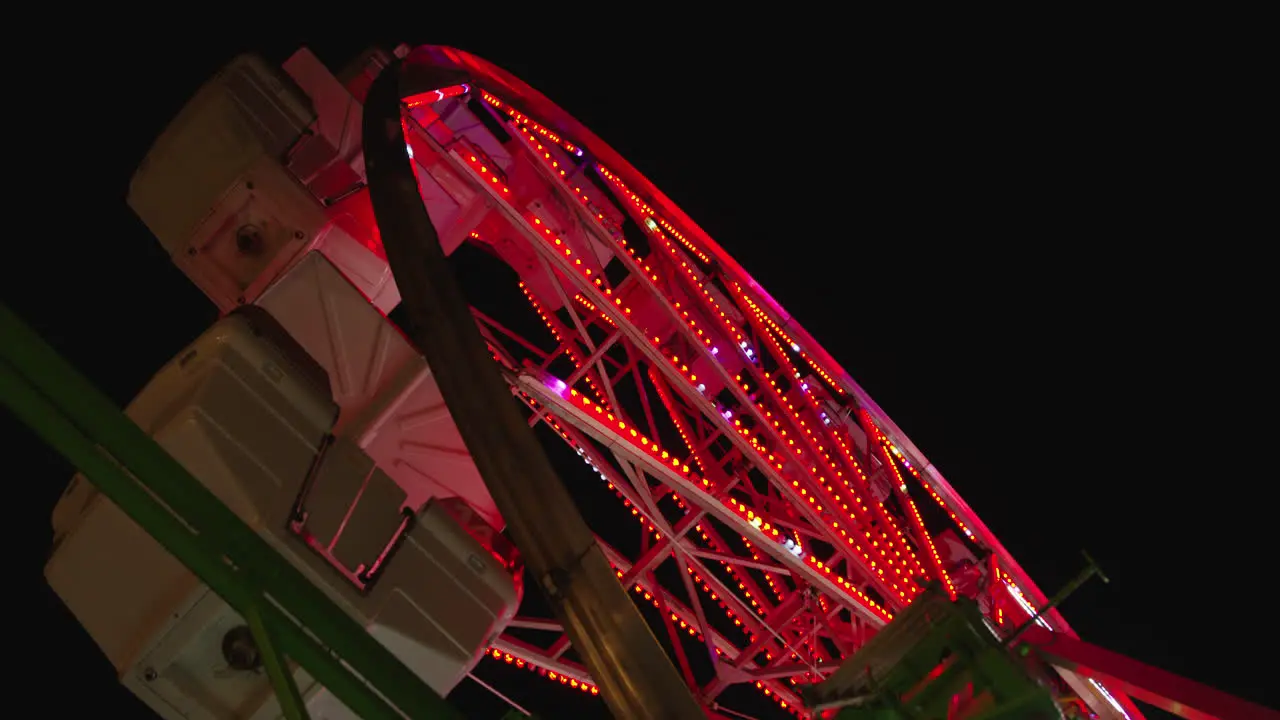 A low angle shot of a Ferris wheel tilts up to reveal the top of it as gondolas swing in the wind at night and the lights of the Ferris wheel change colors