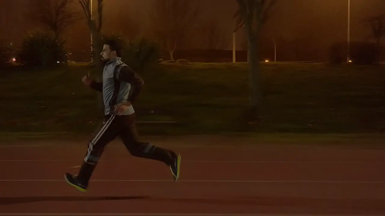 Slow motion video of a young man sprinting on a running track at night while wearing sportswear tracksuit