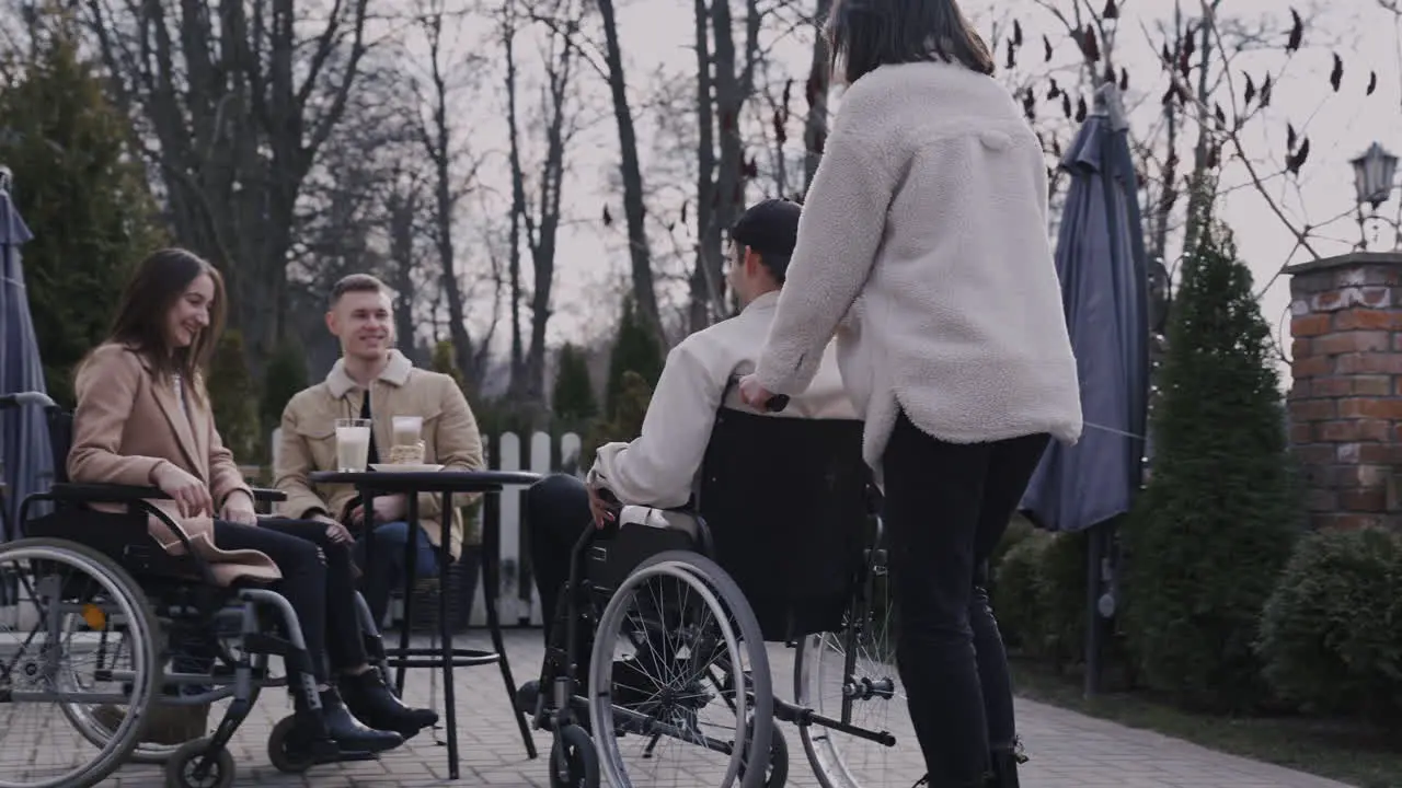 Young Woman And Her Disable Friend In Wheelchair Meeting Other Friends In A Bar Terrace