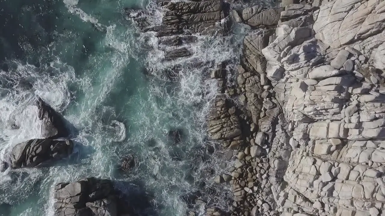 Descending rotating birdseye aerial view above rocky beach splashing rough blue sea waves shoreline