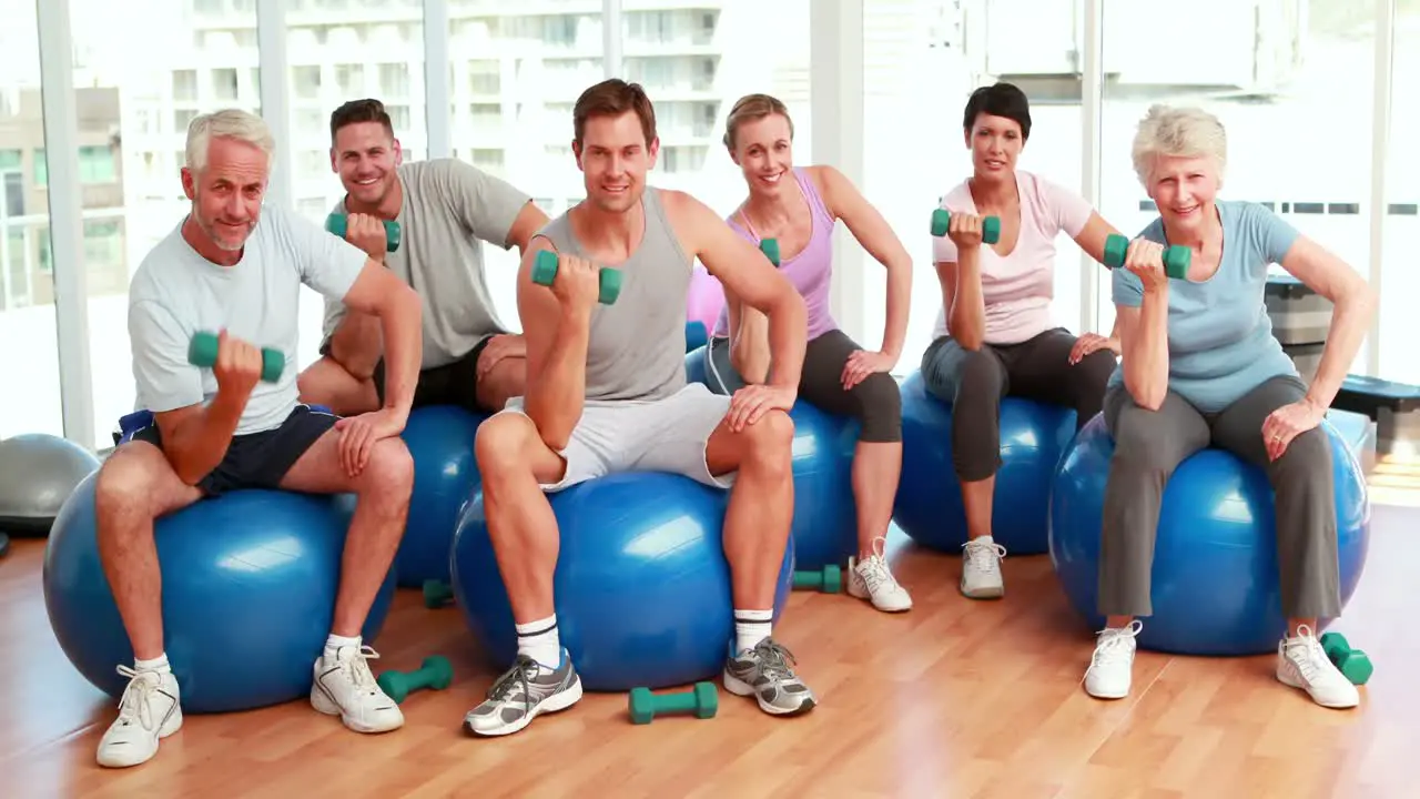 Fitness group sitting on exercise balls lifting hand weights smiling at camera