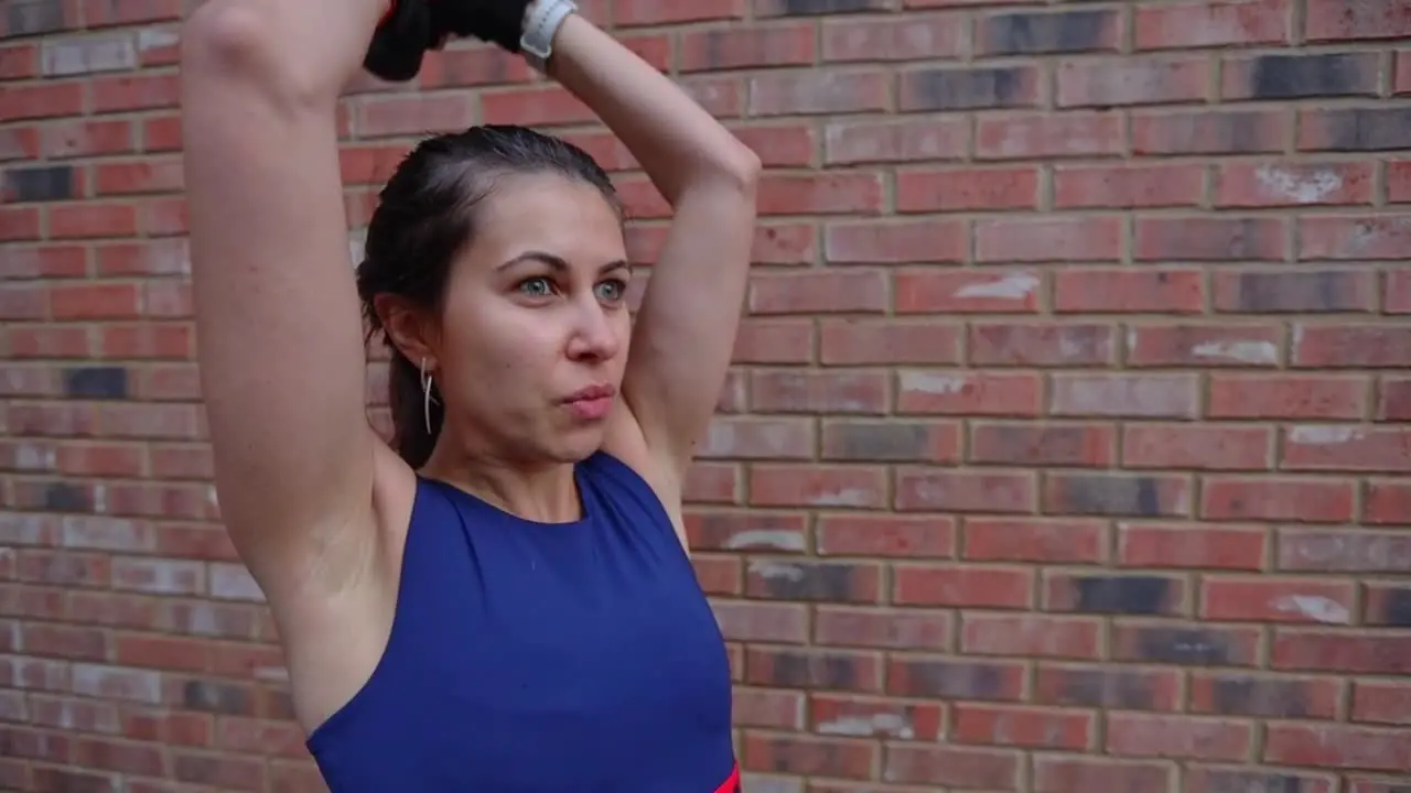 Sporty young Hispanic female raising dumbbell for triceps workout routine next to a brick wall