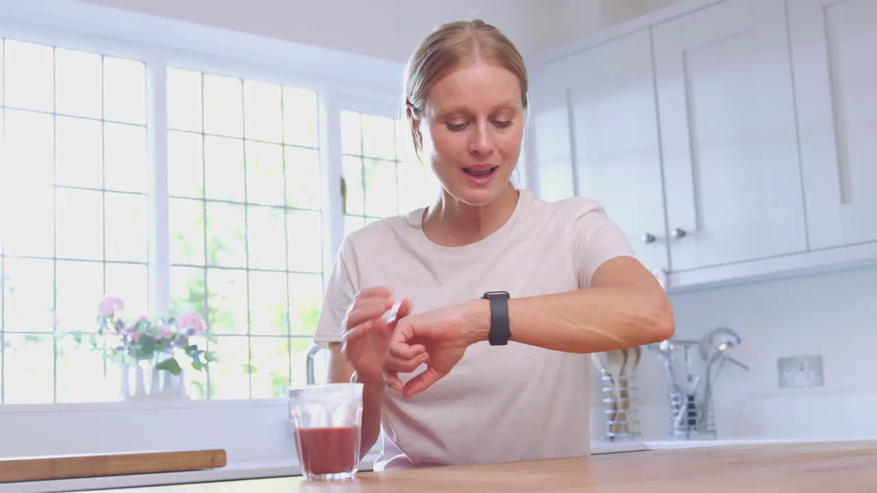 Mature Woman In Fitness Clothing With Health Drink At Home In Kitchen Checks Activity On Smart Watch