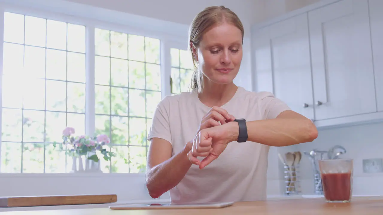 Mature Woman Wearing Fitness Clothing At Home In Kitchen Logging Activity On Digital Tablet