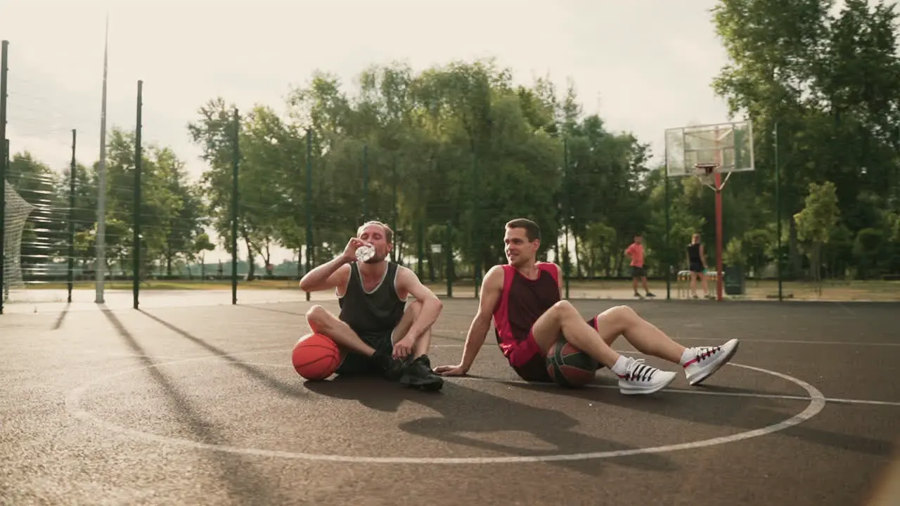 Two Basketball Players Sitting In The Center Of An Outdoor Basketball Court Taking A Break Drinking Water And Talking To Each Other