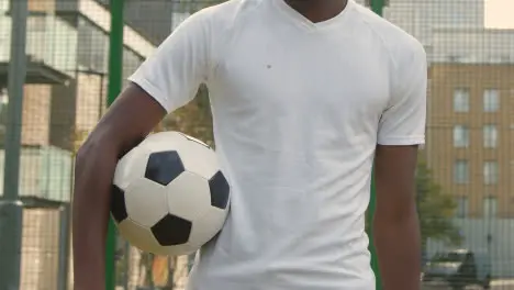 Young Man With Football Under Arm On Artificial Soccer Pitch In Urban City Area 