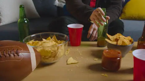 Person At Home Watching American Football Game On TV With Drinks Snacks And Ball