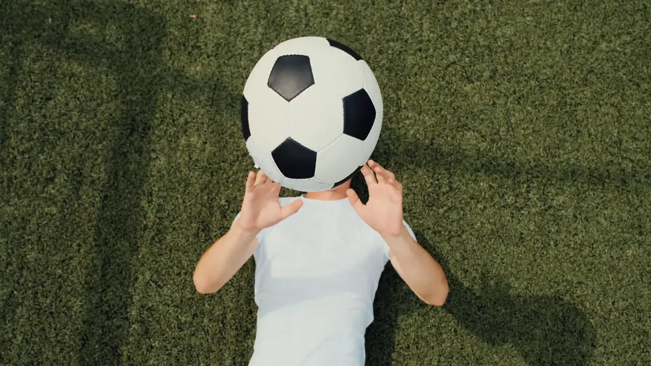 Top View Of A Happy Young Soccer Player Lying On Pitch Throwing And Catching Ball During Training Session