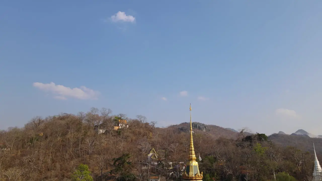 Wat Phra Phutthabat Saraburi Thailand ascending aerial shot of the golden Buddhist temple sliding to the left revealing brown trees in summer a hill with a stairway going up to another temple