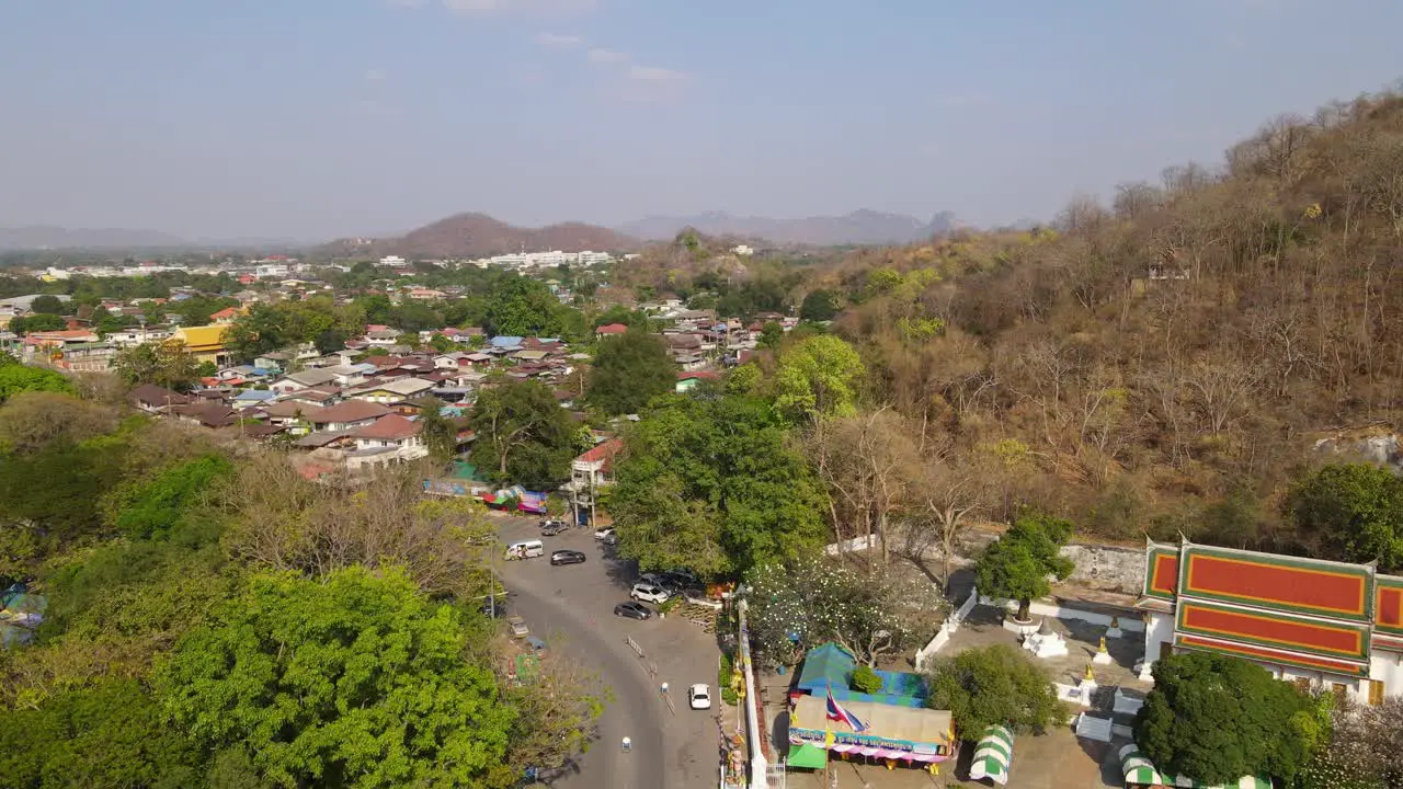 Wat Phra Phutthabat Saraburi Thailand ascending aerial footage revealing the road with cars Thai Flag entrance into the teple community and houses hills and the horizon blistering summer day