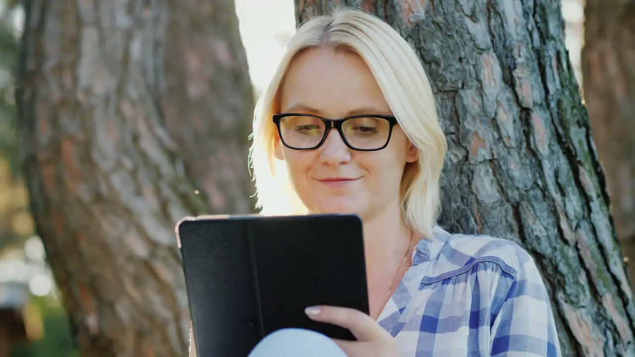 Attractive Young Woman In Glasses Uses A Tablet Sits In A Park Near A Tree Beautiful Light Before Su