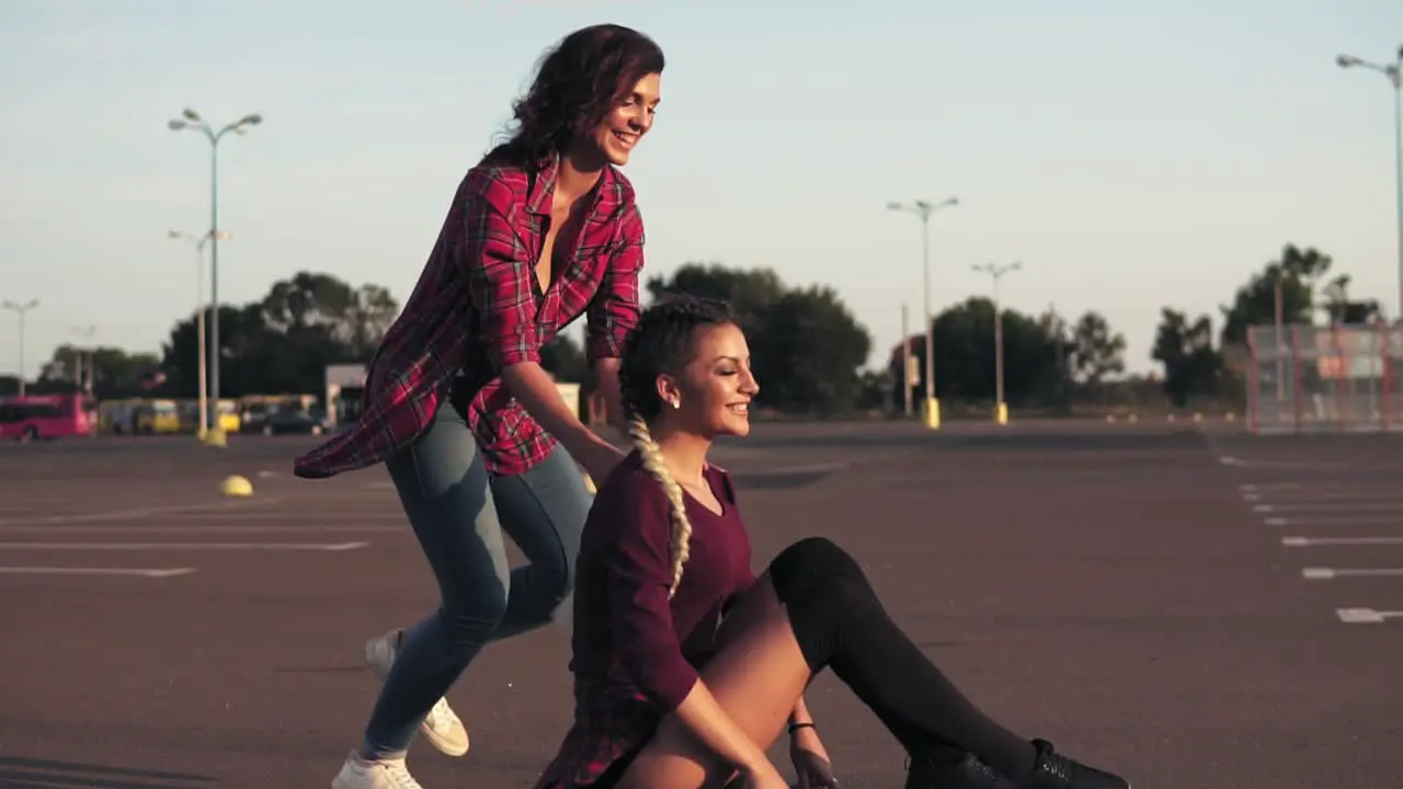 Smiling Woman Sitting On A Longboard While Her Friend Is Pushing Her Behind And Running During Sunset