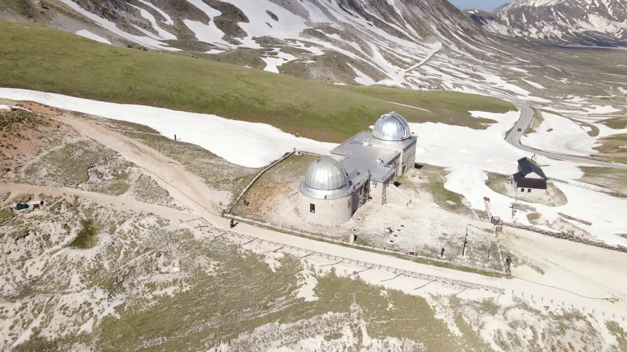 Wide angle drone shot panning upwards of a busy observatory with hikers walking by revealing the mountain landscape in the distance in the region of Abruzzo in Italy