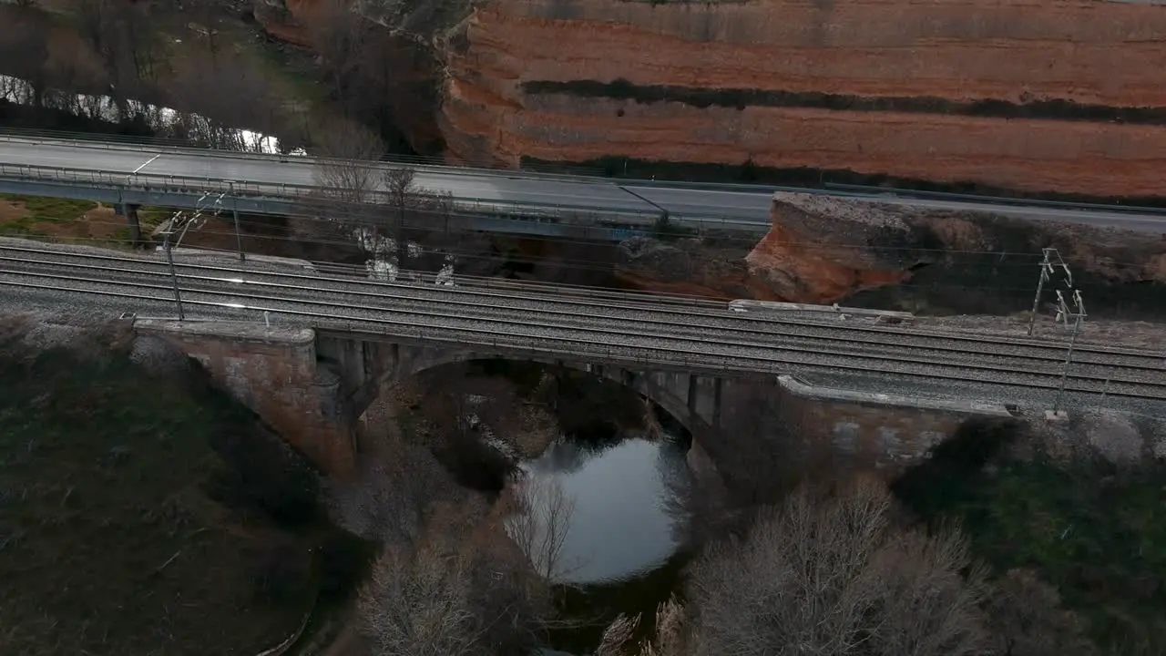 Aerial shot moving backwards from a railway over a river with a train passing through it near a traffic road and a dirt road