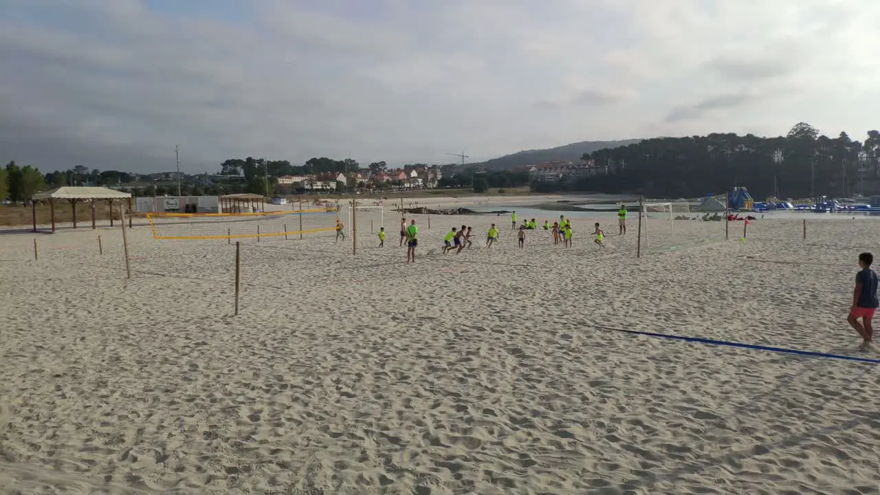 Adult beach soccer coach with kids watches the activity among the volleyball nets in flashy colored T-shirts one summer morning rolling shot to the left