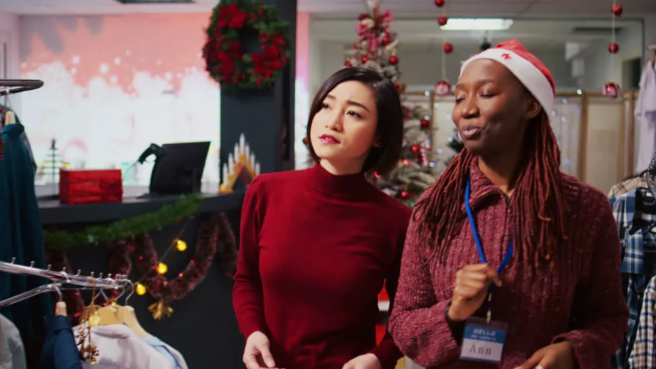 Tracking shot of helpful retail assistant in festive decorated clothing store showing asian customer beautiful red garments ready to be worn at Christmas themed office party