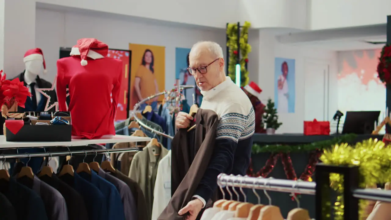 Dolly out shot of elderly client in xmas decorated shopping mall store trying to determine if stylish blazer is fitting him Senior shopper in fashion boutique during Christmas sales