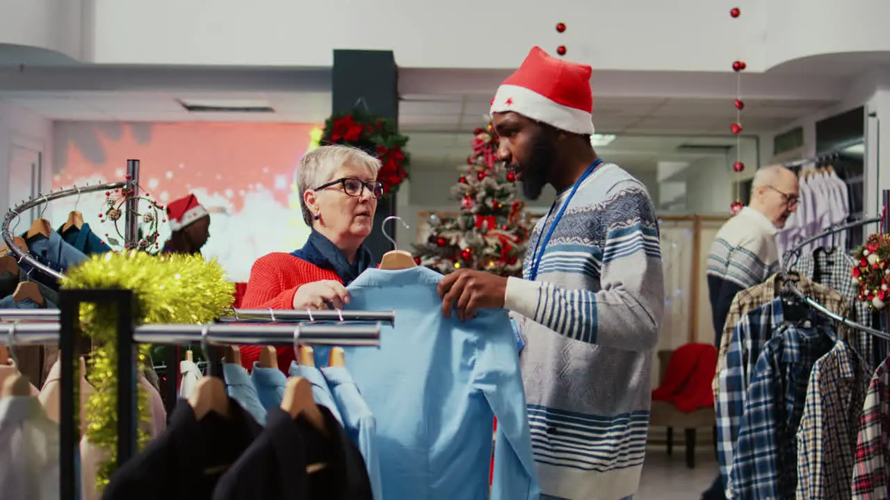 Aged customer browsing through clothes in festive xmas ornate shopping mall fashion store men section being assited by employee with finding shirt to gift husband for upcoming winter holiday season