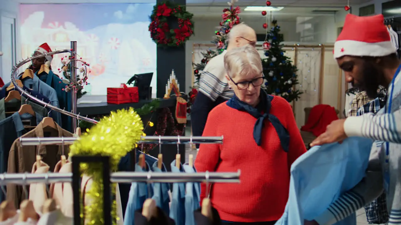 Worker assisting senior customer with picking right outfit to give as present to husband for upcoming xmas holiday season Client browsing through clothes in festive ornate fashion shop