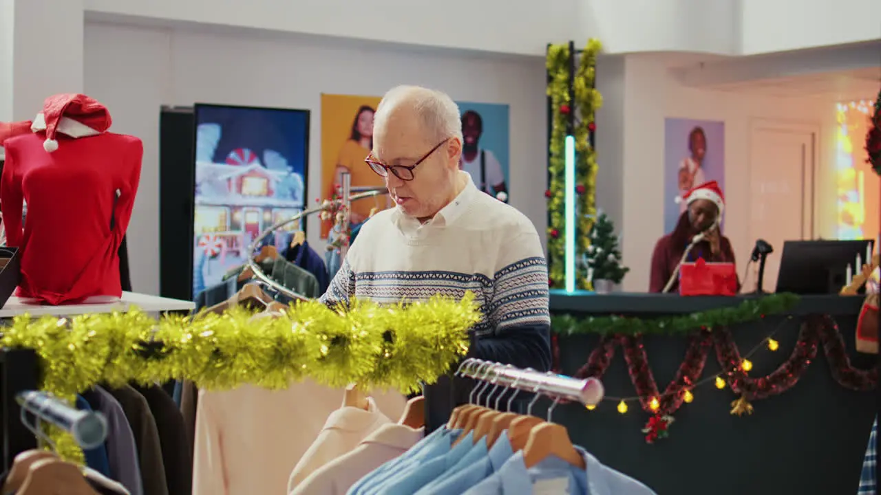 Dolly in shot of senior customer in festive ornate shopping mall store trying to determine if stylish blazer is fitting him Aged client in fashion shop during Christmas holiday season