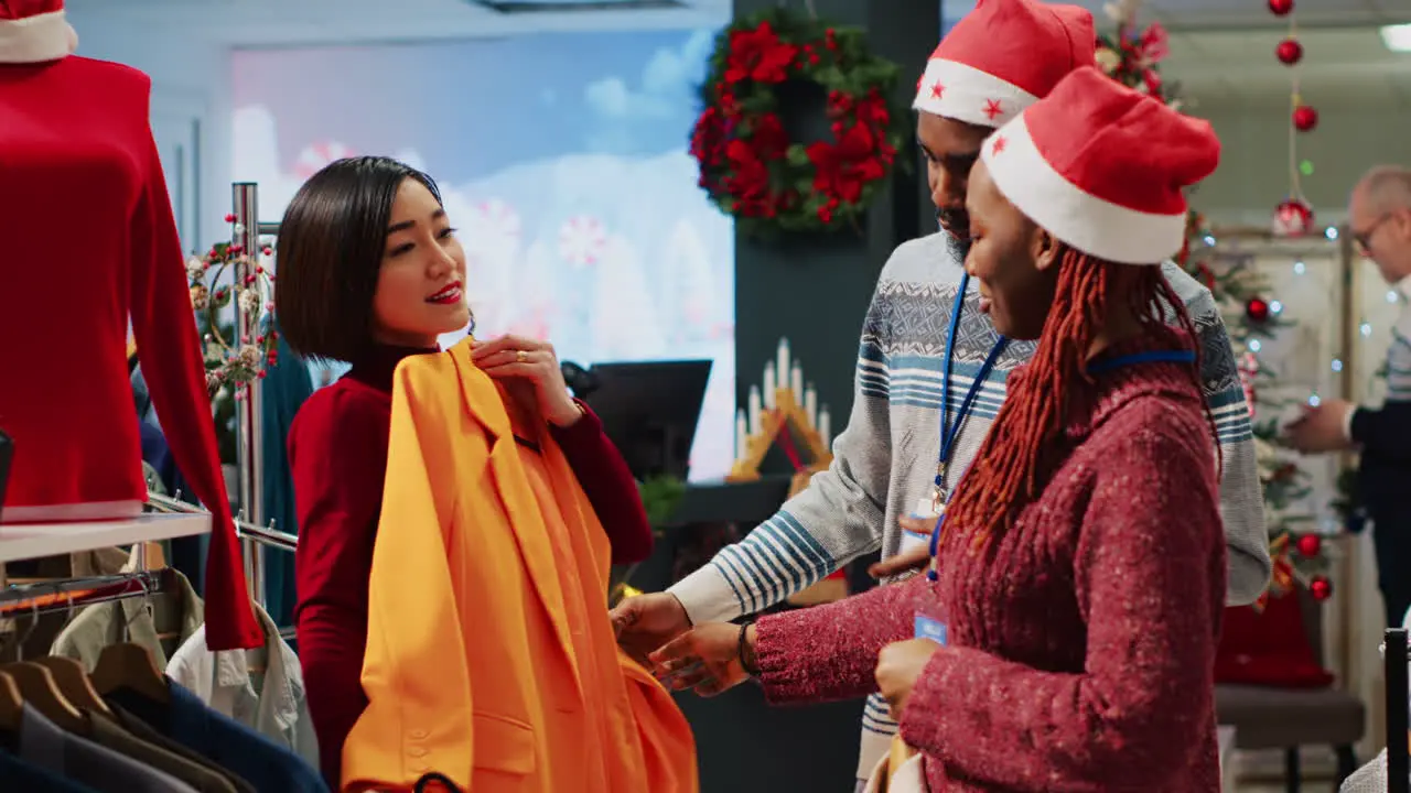 Asian retail assistant and coworker helping indecisive customer choose between two colors of blazers in Christmas decorated clothing shop Employees assisting client during winter holiday season