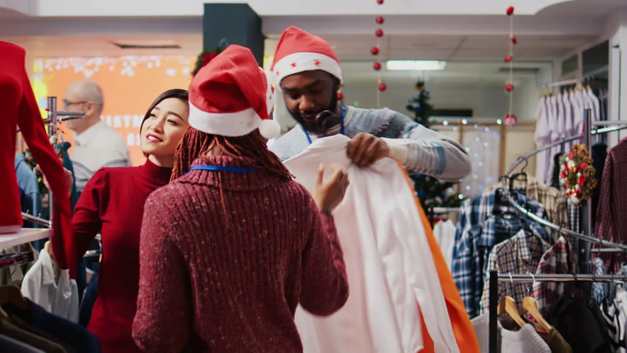 Friendly retail assistant in festive ornate shopping mall fashion boutique showing client beautiful red garments while african american coworker brings stylish blazers for her to try on