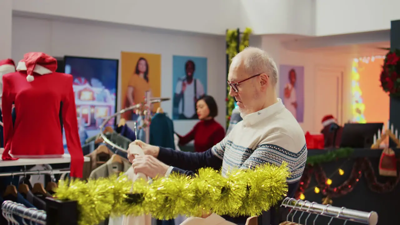 Dolly out shot of senescent man in festive decorated clothing store trying to determine if luxurious blazer is the right fit Older client in Christmas adorn fashion shop during holiday sales