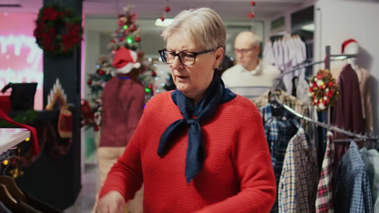 Elderly woman browsing through clothes in festive Christmas decorated shopping mall fashion shop men section looking for blazer to gift husband for upcoming xmas holiday season