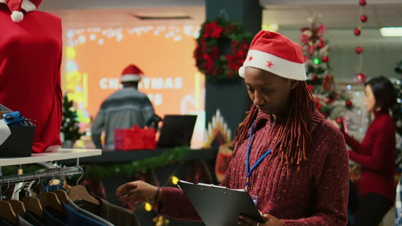 Portrait of smiling employee wearing Santa hat walking through clothing rack rows in xmas adorn shopping mall store using clipboard to crosscheck price tags making sure shop is ready for customers