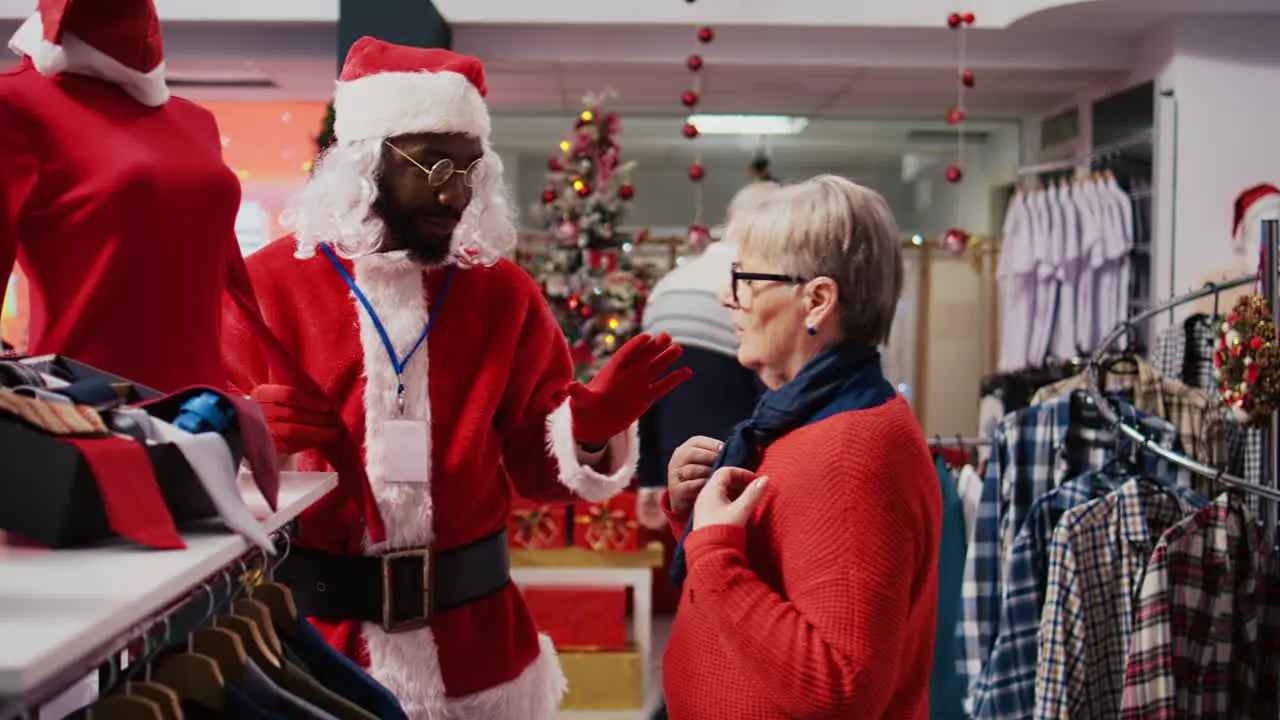 Manager wearing Santa Claus costume around shopping mall fashion boutique chatting with elderly woman Supervisor in holiday themed suit showing senior client red garment piece