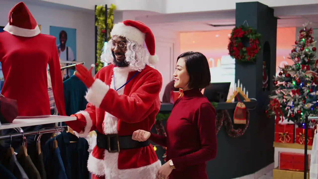 Manager wearing Santa Claus costume around shopping mall fashion boutique to spread holiday joy Supervisor in festive themed suit showing cheerful client red garment piece