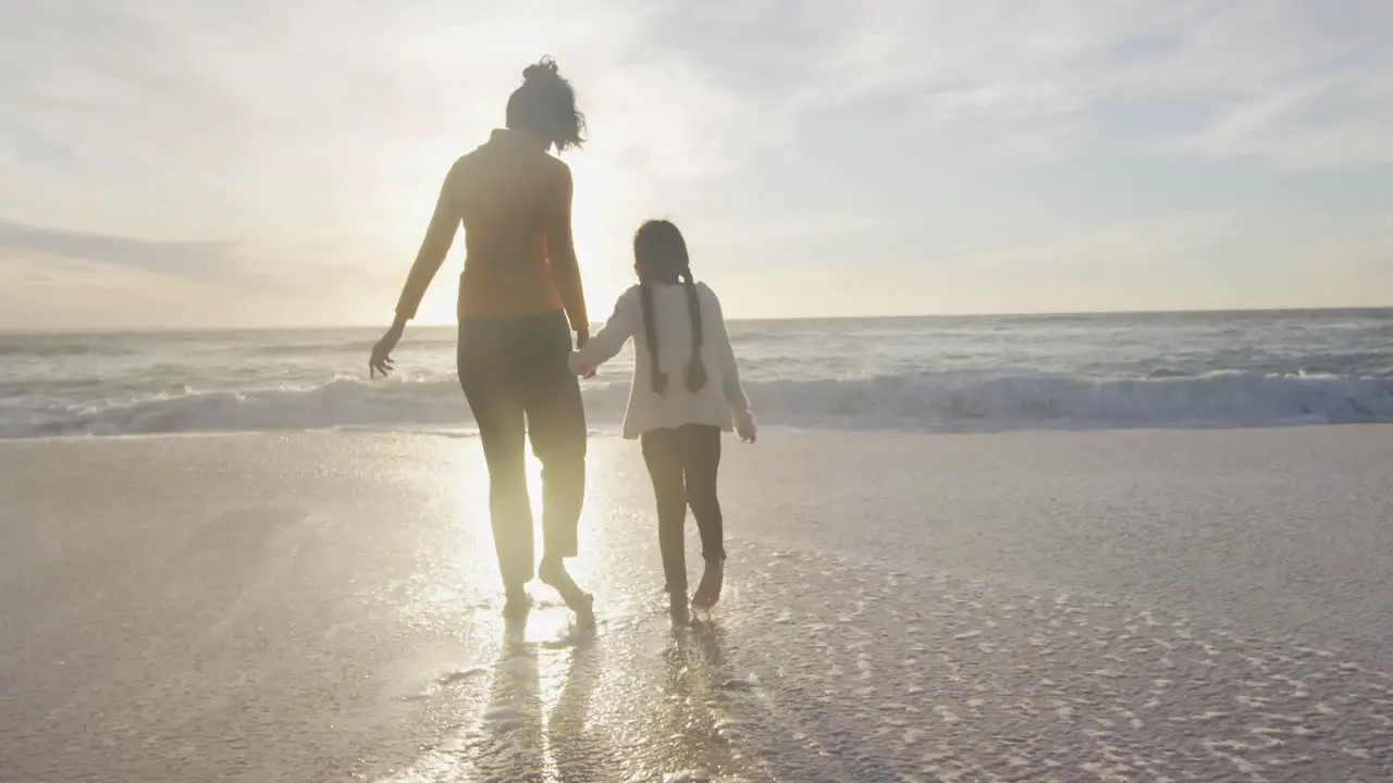 Back view of hispanic mother and daughter walking on beach at sunset