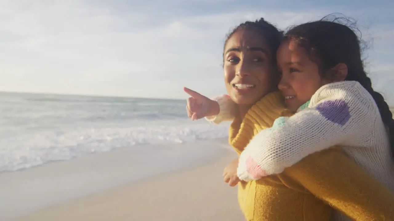 Happy hispanic mother carrying piggyback daughter and looking at sunset on beach