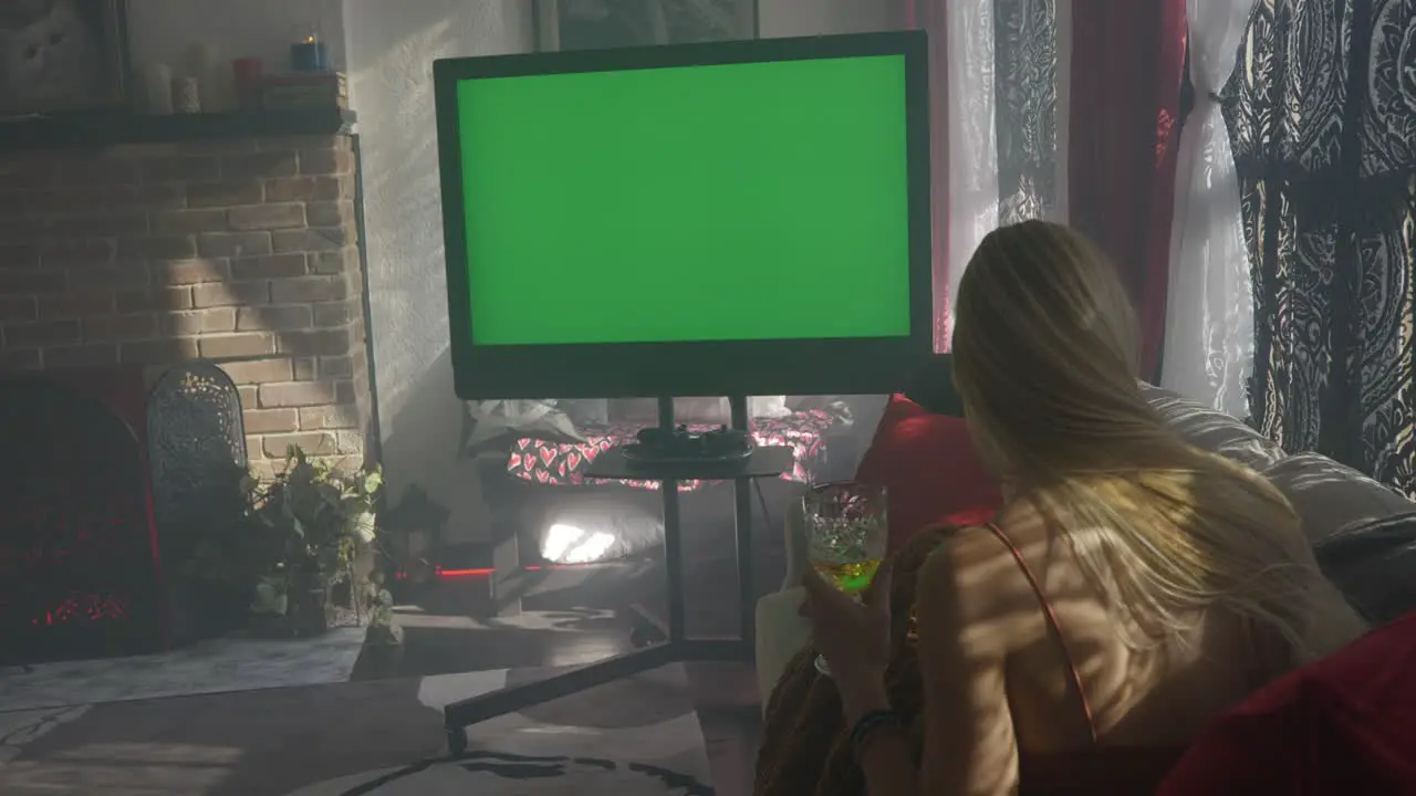 Green screen television on misty hazy morning with blonde women lady girl sitting on couch cuddled up with blanket as someone hands her champagne wine glass as she watches the tv