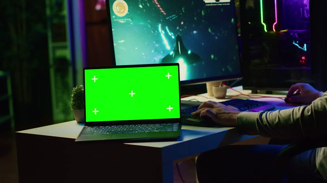 Green screen laptop next to man having fun by using gaming keyboard to fly spaceship in SF videogame