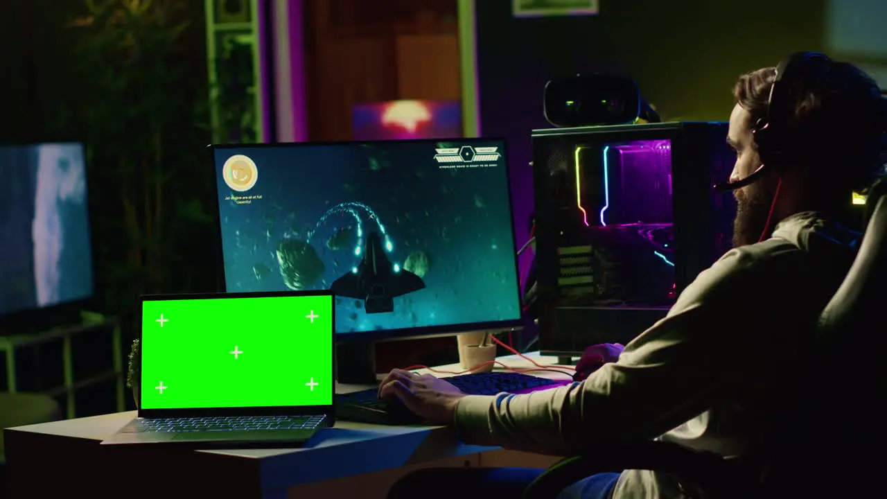 Green screen laptop next to man using gaming keyboard to play spaceship flying singleplayer game