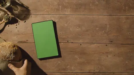 Overhead Shot Of Person Picking Up Loaf Of Bread From Wooden Table With Green Screen Digital Tablet