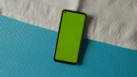 Overhead Studio Fitness Shot Of Rotating Green Screen Mobile Phone With Trainers And Towel On Blue Exercise Mat