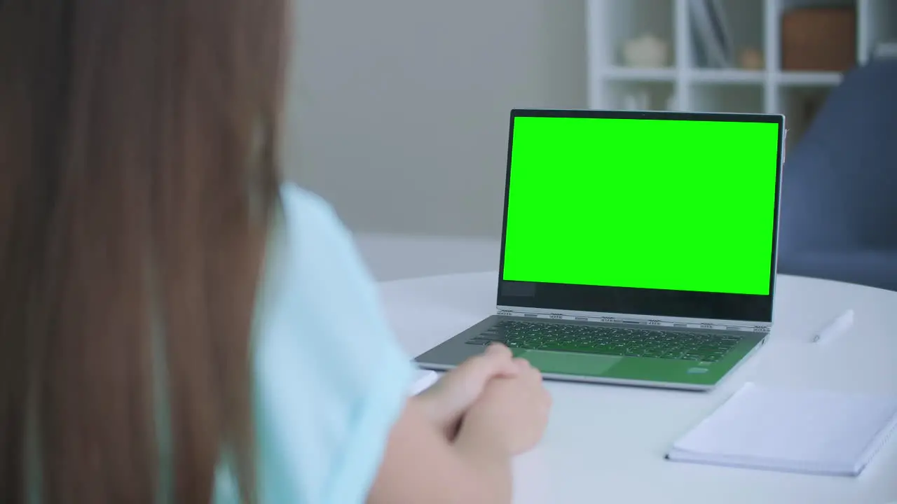 Woman sits at desk in bedroom she looks at laptop green screen and talks to someone over internet video communications sometimes taking notes in notebook Close-up