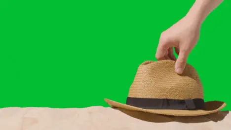 Summer Holiday Concept With Hand Picking Up Sun Hat On Sand Against Green Screen