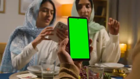 Man Looking At Green Screen Mobile Phone Sitting Around Muslim Family Table At Home Eating Iftar Meal Breaking Daily Fast During Ramadan