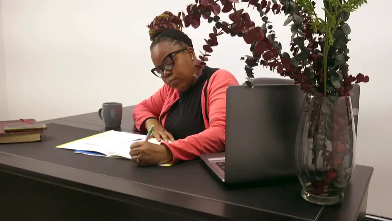 Black business woman wearing glasses writing at a desk in office dressed in business attire