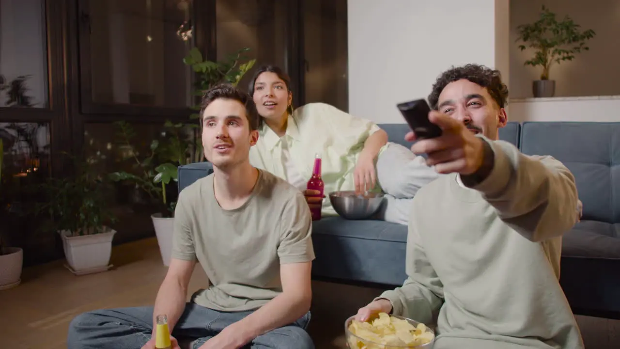 Three Friends Watching Comic Movie On Television Sitting On Couch And Floor Eating Chips And Drinking Soda