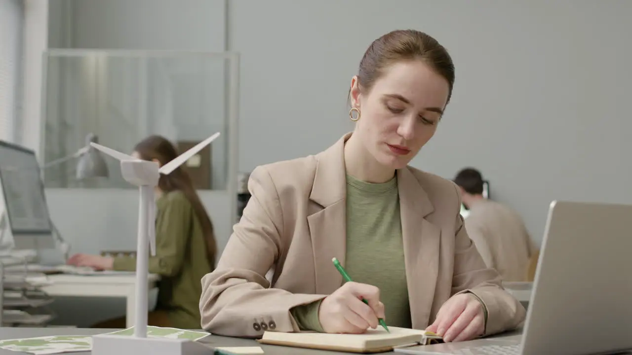 Woman Using Laptop And Writting Notes Sitting At Table With Windmill Model In The Office