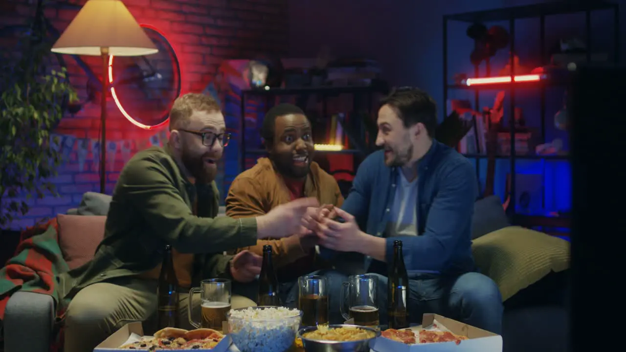 Guy Coming Into The Dark Living Room To His Mixed Races Friends Sport Fans Sitting On The Couch Doing Cheers Gesture With Beer While Watching Match And Cheering Up